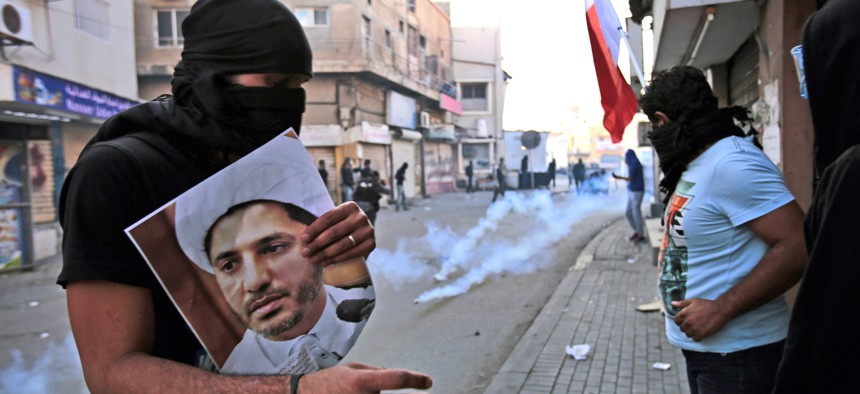 In this Jan. 3, 2015, file photo, a masked Bahraini anti-government protester holds a picture of jailed Shiite cleric Sheik Ali Salman, the head of the opposition al-Wefaq political association, as riot police fire tear gas canisters during clashes in Bil